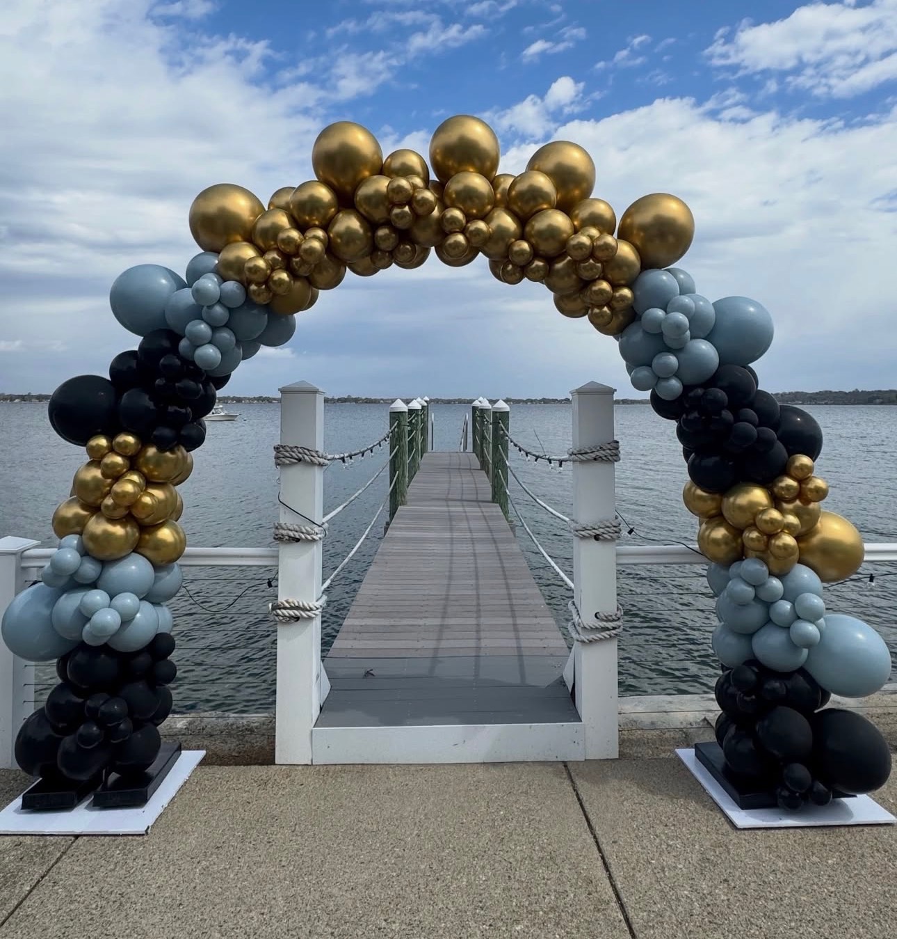 Waterfront balloon arch in gold, dusty blue and black over a dock