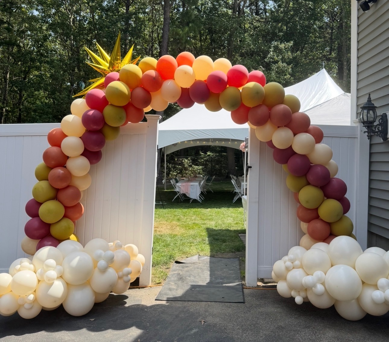 Sunset-toned spiral balloon arch with gold starburst over a backyard tent entry